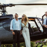 Elegantný pár stojaci pri helikoptére po ukončení vyhliadkového letu, usmievajúci sa vedľa pilota na malebnom pozadí.